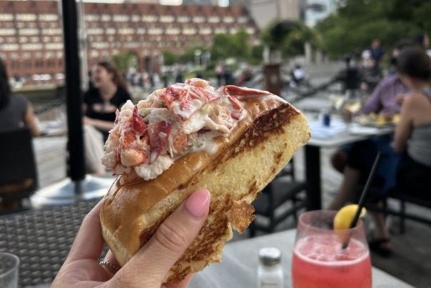 Hand holding a lobster roll, a pink cocktail next to it, and blurred background of people eating outside