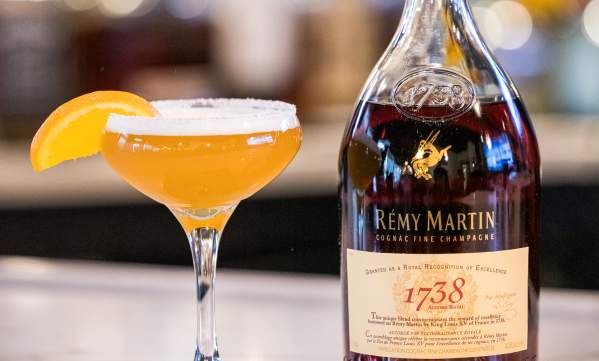 Cocktail glass with orange drink, orange segment, and bottle of cognac on a bar counter