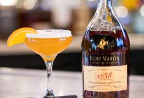 Cocktail glass with orange drink, orange segment, and bottle of cognac on a bar counter