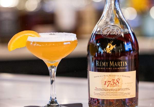 Cocktail glass with orange drink, orange segment, and bottle of cognac on a bar counter
