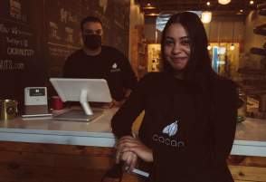 Woman leaning on counter in front of man wearing a cloth mask working behind a register at Cacao